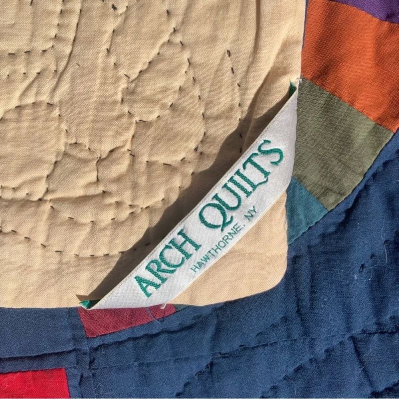 A close-up of a colorful quilt shows a fabric label reading ARCH QUILTS HAWTHORNE, NY sewn into the seam, with visible stitch patterns and various fabric colors in the background.