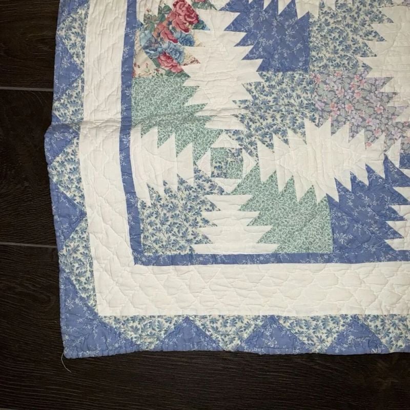 A close-up of a quilt corner featuring a blue, green, and white patchwork pattern with floral designs and geometric shapes, bordered by a blue and white floral print fabric, laid on a dark wooden floor.
