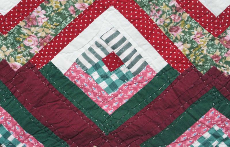 A close-up of a colorful patchwork quilt by Arch Quilts, featuring geometric diamond patterns with red, green, white, and floral fabric strips sewn together in a symmetrical, repeating design.