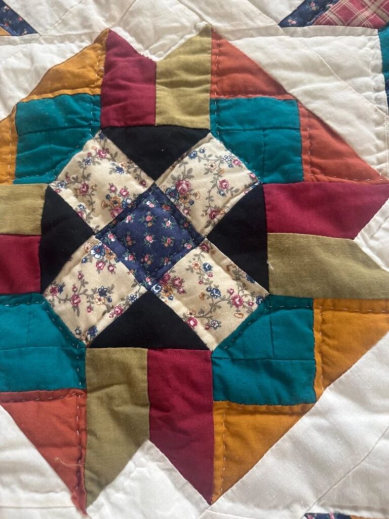 A close-up of an Arch Quilts creation, showcasing a colorful quilt with a geometric star pattern, featuring teal, mustard, rust, black, cream, and floral print fabrics sewn together with visible stitching.