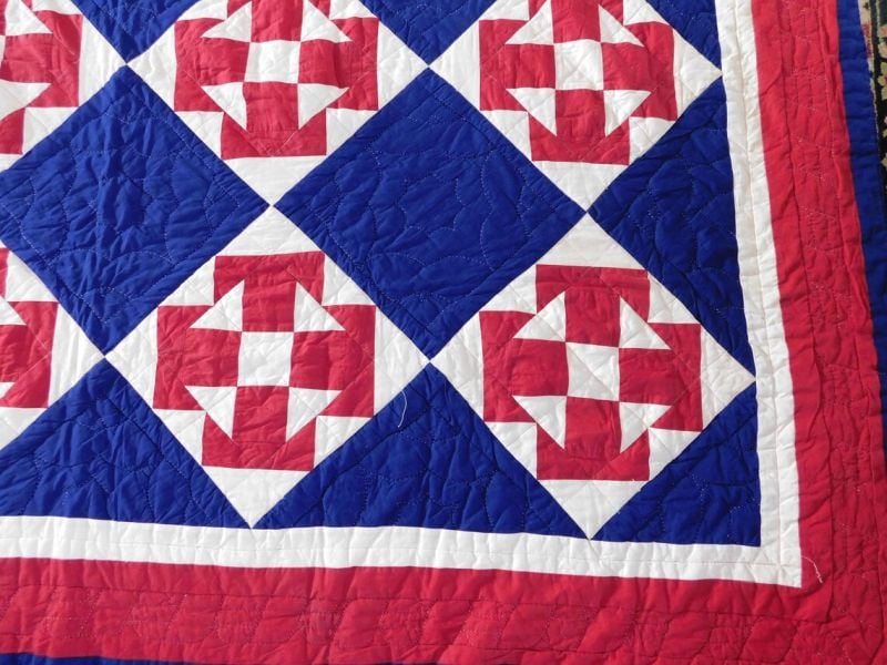 An Arch Quilts design, this quilt features a geometric pattern of red and white shapes on a blue background, bordered by white and red strips. The repeating diamond and triangle motifs add to its striking visual appeal.