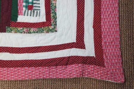 A close-up of a quilt corner featuring patterned fabric strips in red, white, green, and pink with floral and polka dot designs, lying on a textured brown carpet.