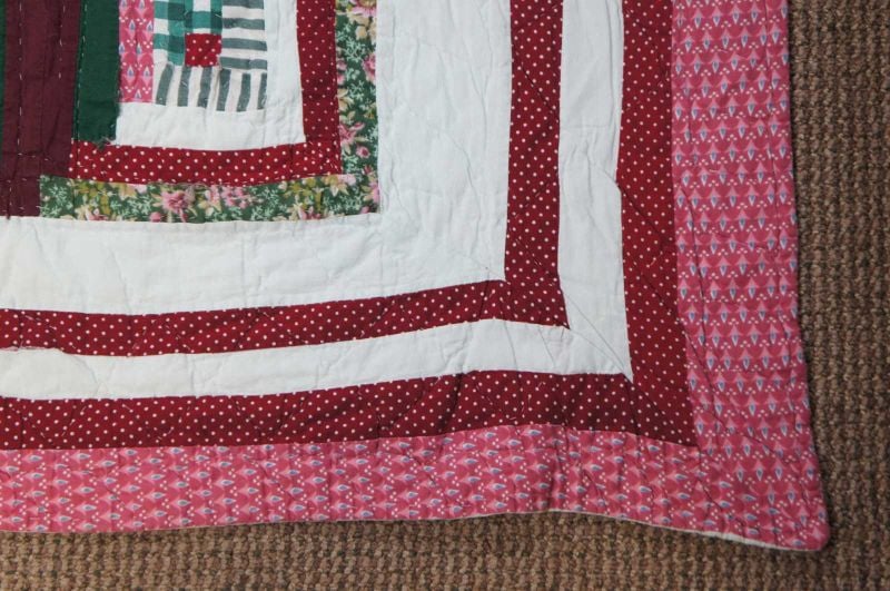 A close-up of a quilt corner featuring patterned fabric strips in red, white, green, and pink with floral and polka dot designs, lying on a textured brown carpet.