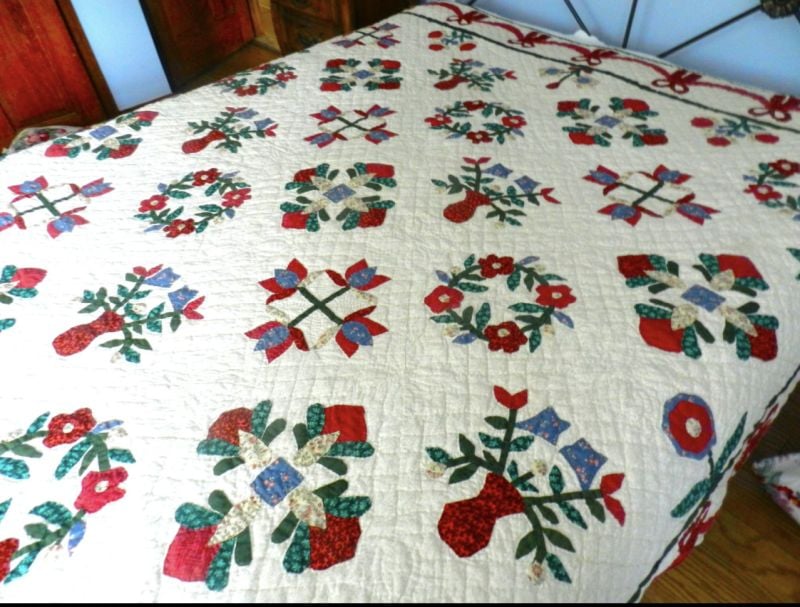 Arch-quilt-from-facebook-marketplace A handmade quilt covers a bed, featuring colorful floral and geometric patchwork patterns in red, green, blue, and cream tones. The quilt displays a repeating design with flower motifs and leaves.