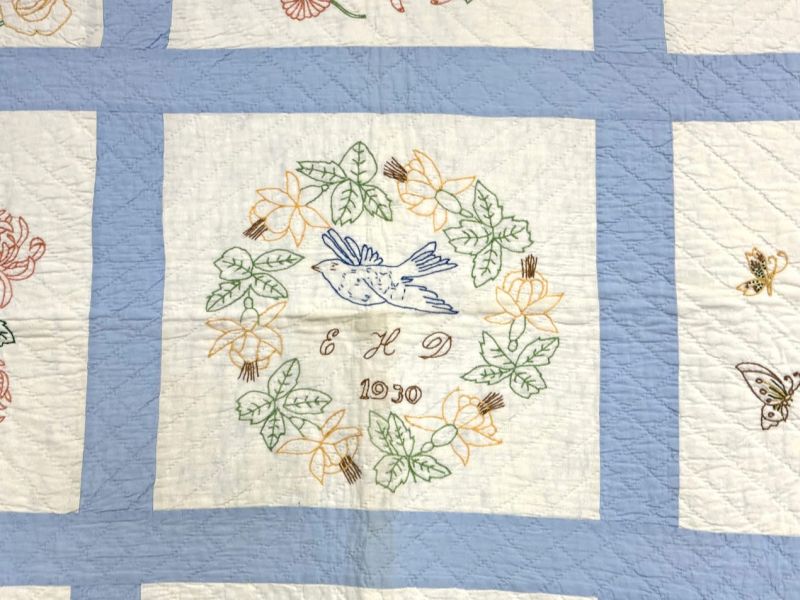 A quilt square featuring an embroidered blue bird surrounded by a floral wreath, with the initials ELD and the year 1930 stitched beneath. The background is white with a blue border.