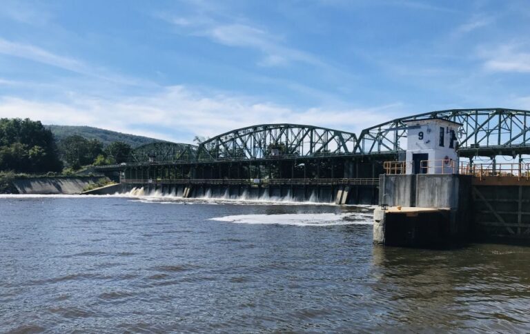 A green metal bridge with a lock and dam structure spans a wide river under a blue sky, with water flowing over the dam and trees visible on the left—echoing stories of Underground Railroad Quilts that guided many to freedom.