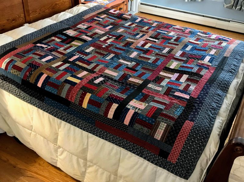 A colorful handmade quilt with various geometric patterns and multicolored fabric pieces lays neatly on a bed with a white comforter in a cozy, sunlit bedroom.