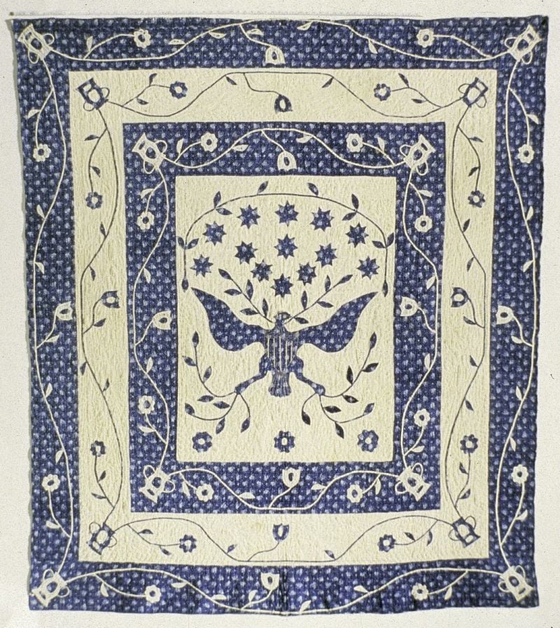 The Great Seal Quilt A square quilt with a blue and white floral pattern featuring a central eagle with a basket of flowers, surrounded by vine and flower motif borders—an elegant piece reminiscent of styles seen in The Smithsonian Quilt Controversy.