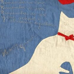 A piece of fabric art features a white cat with a red bow on a blue background, and hand-stitched text that reads: Remember us now, Remember us ever, Remember us as your school children together. Harry Ygeant.