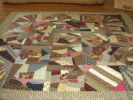 A handmade quilt with a patchwork design featuring various colorful fabric scraps in different shapes and patterns, bordered by a patterned edge, is spread out on a wooden floor.