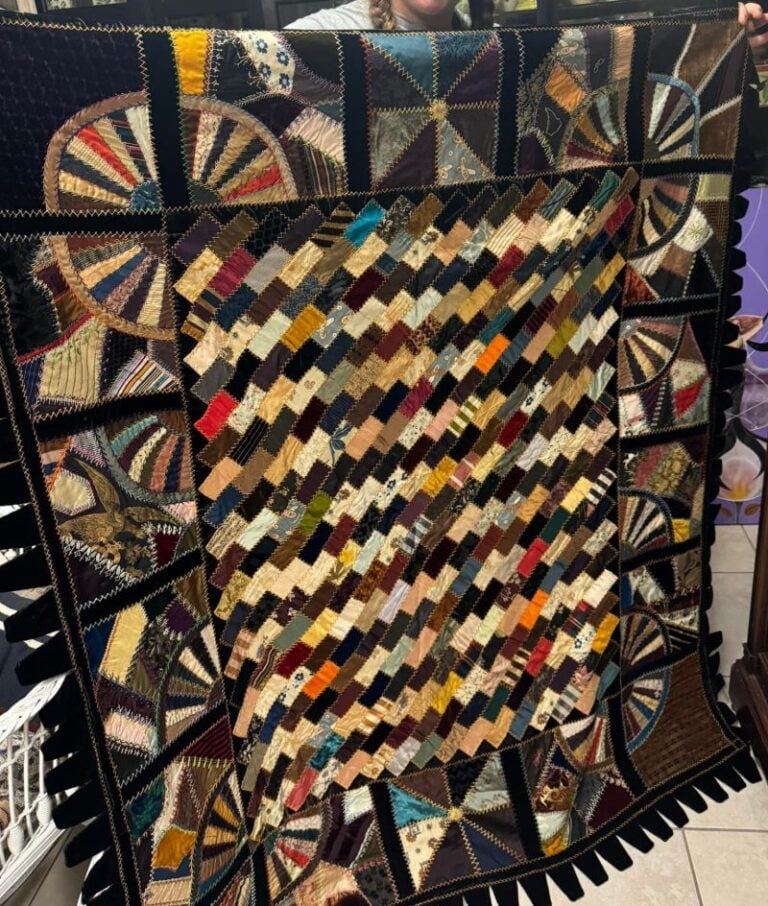 crazy-beauty A handmade quilt with a colorful patchwork diamond pattern in the center, bordered by fan-shaped patchwork sections and black zigzag edging, held up indoors by an unseen person.