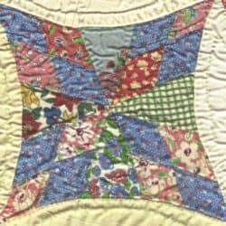 A colorful quilt block with various floral and geometric patterned fabric pieces in blue, pink, green, and white, sewn together in an abstract design on a light quilted background.