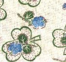 A fabric pattern featuring green four-leaf clovers with small blue clusters in the center, set against a light, textured background. The design is simple and has a hand-drawn, vintage feel.