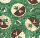 A green textured fabric with a repeating pattern of circles, each divided in half with contrasting dark and light sections, featuring abstract leaf and triangle shapes inside. Smaller outline circles are scattered between the larger ones.