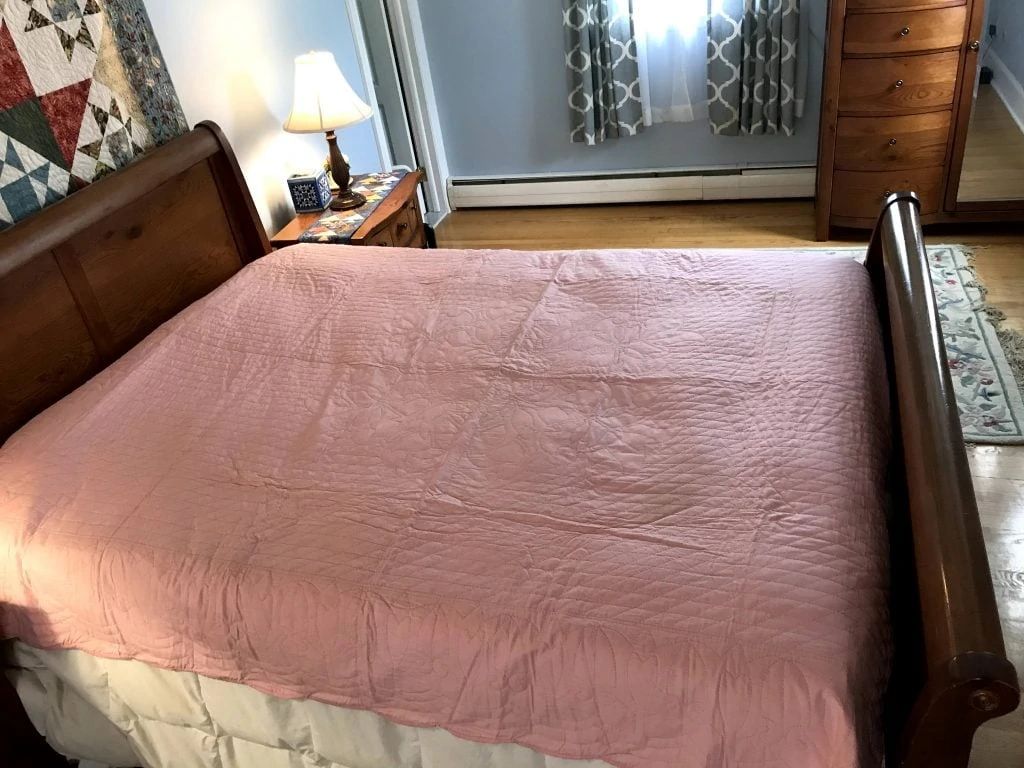 A neatly made bed with a light pink quilt in a cozy bedroom. There is a wooden headboard and footboard, a bedside table with a lamp, a dresser with a mirror, and a window with patterned curtains.