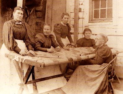 Four women in old-fashioned clothing sit and stand around a large wooden quilting frame on a porch, working together on a quilt. The scene appears to be from the late 19th or early 20th century.