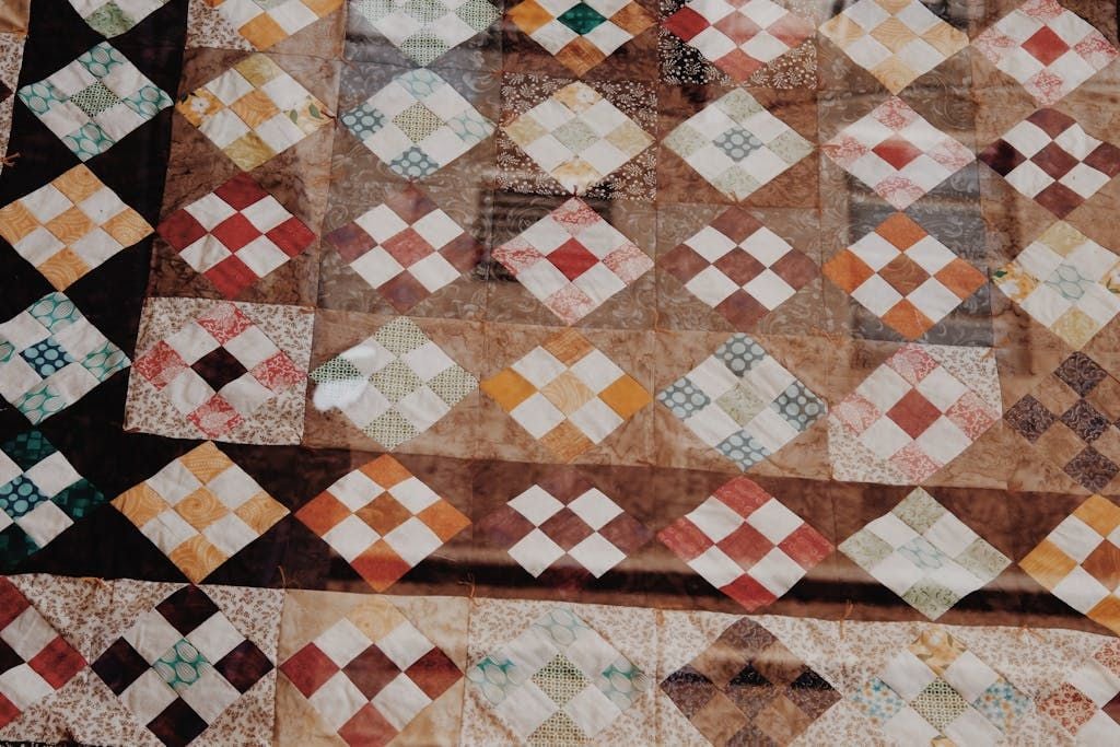 A handmade quilt featuring a patchwork pattern of colorful diamond and square shapes in earthy tones like brown, red, cream, and green, arranged in a repeating geometric design.