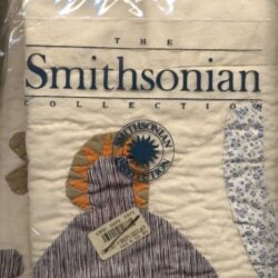 A quilt with an applique design and blue flower pattern is packaged in plastic with The Smithsonian Collection and a circular Smithsonian logo printed on it, referencing The Smithsonian Quilt Controversy. A price sticker is attached to the package.