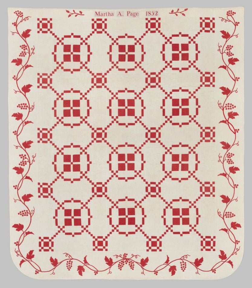 Burgoyne-Surrounded A quilt with a red and white geometric pattern, featuring circular and square motifs inspired by the Burgoyne Surrounded quilt block myth. The border has red vines and leaves. “Martha A. Page 1852” is stitched at the top center.
