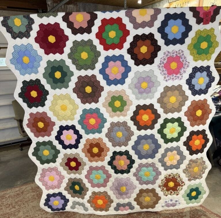 Grandmothers Flower Garden Quilt A handmade quilt with a white background featuring rows of multicolored hexagon flower patterns in various fabrics, displayed indoors.