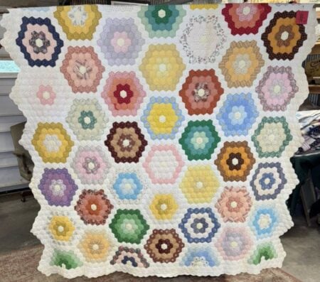 Grandmas-Flower-Garden A handmade quilt featuring a pattern of colorful hexagon flower shapes on a white background, each flower made of concentric rings of fabric in various pastel and bright colors. The edges have a scalloped appearance.