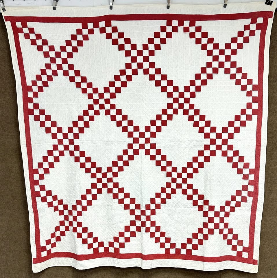 Irish-Chain-red A handmade quilt featuring a repeating diamond pattern made of small red and white squares, bordered by a red and white striped edge, hangs on a wall.