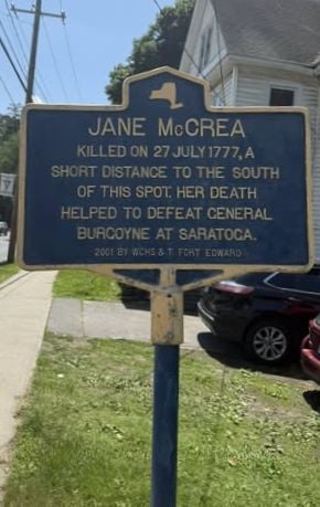 Jane-McCreaA blue and y ellow historical marker sign describes Jane McCrea, killed on July 27, 1777, noting her death helped defeat General Burgoyne at Saratoga. The sign is on a sidewalk near a street, house, and parked cars.