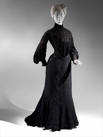 Mourning-Dress-with-black-silk-crepe A mannequin wears a long, black, high-necked Victorian-style dress with lace details, long sleeves, and a fitted waist, standing in a dramatic pose against a plain, gradient background.