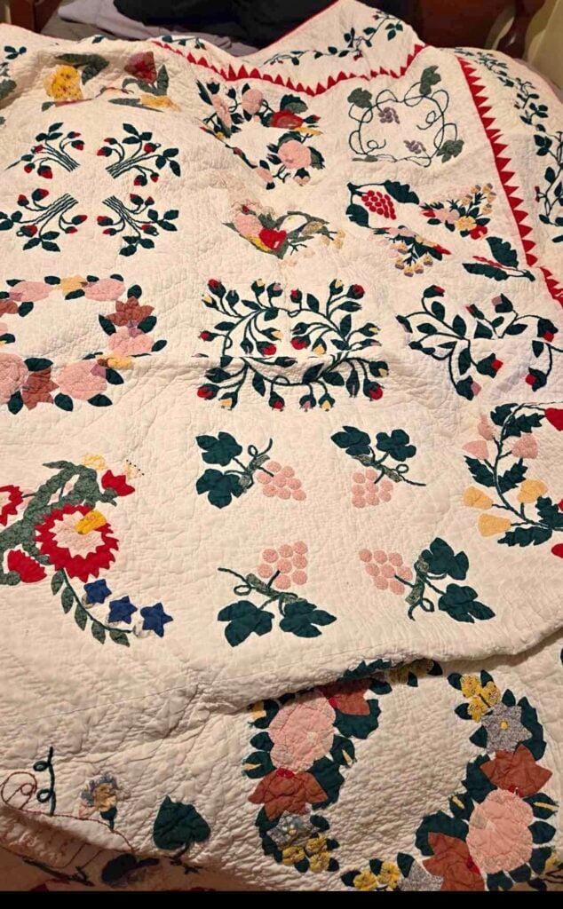 A colorful handmade quilt with intricate floral and leaf patterns in green, red, yellow, pink, and blue, spread out over a bed. The quilt has a white background with a red triangular border along one edge.