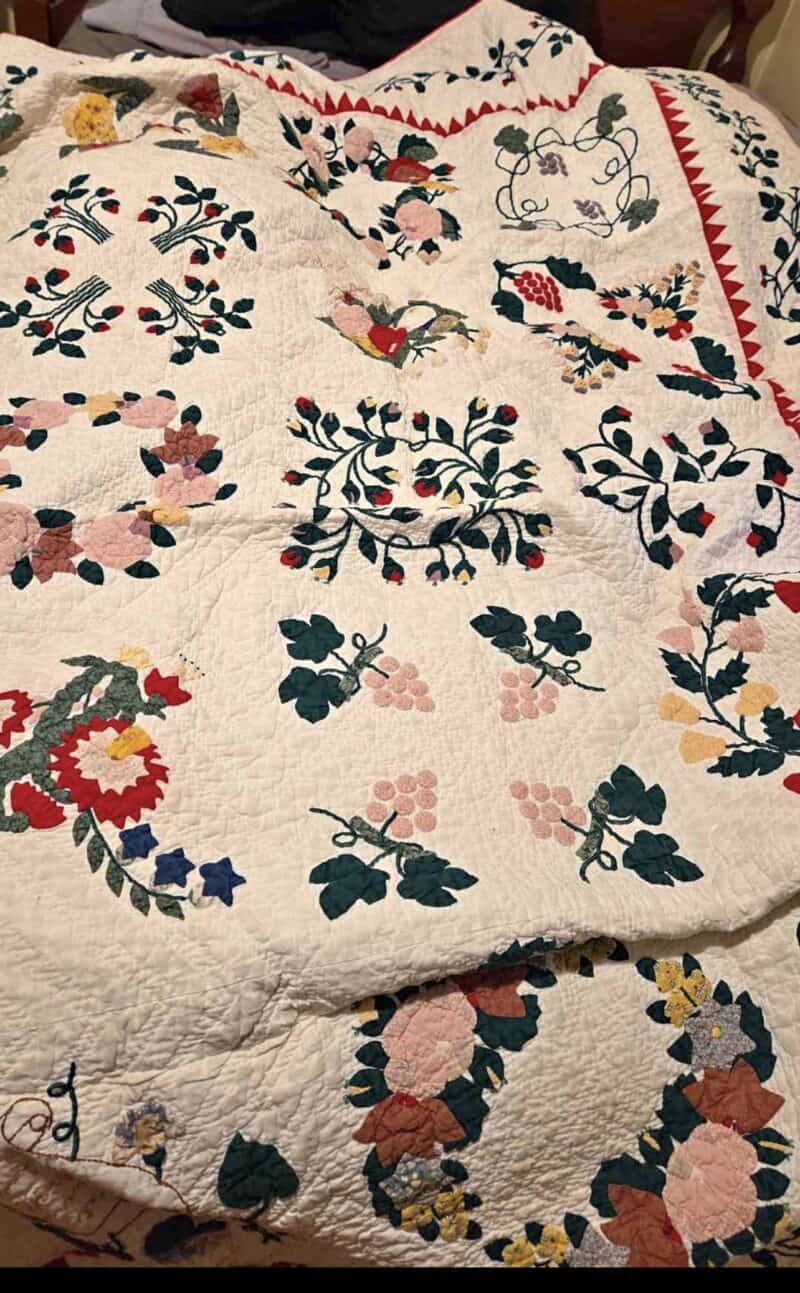 A colorful handmade quilt with intricate floral and leaf patterns in green, red, yellow, pink, and blue, spread out over a bed. The quilt has a white background with a red triangular border along one edge.