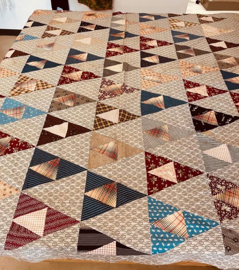 Th ousand-Pyramid-QuiltA handmade quilt displayed on a bed features rows of triangle patches in various colors and patterns, including blue, red, brown, and beige, set against a light patterned background.