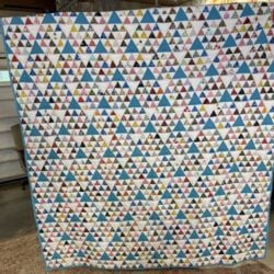 A large handmade quilt featuring a repeating pattern of colorful, small triangles arranged in rows. The triangles include various colors and prints, bordered by a light blue edge. The quilt is displayed indoors.