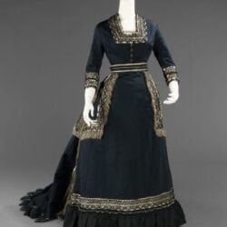 Victorian-Mourning-Featured-Image Dark blue Victorian-style dress inspired by Victorian Mourning Customs in the United States, with long sleeves, square neckline, ornate gold trim, fitted bodice, flared skirt and train, plus decorative fringe. Displayed on a mannequin against a gray background.