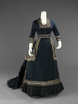 Victorian-Mourning-Featured-Image Dark blue Victorian-style dress inspired by Victorian Mourning Customs in the United States, with long sleeves, square neckline, ornate gold trim, fitted bodice, flared skirt and train, plus decorative fringe. Displayed on a mannequin against a gray background.