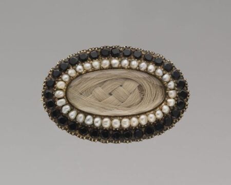 An oval brooch featuring a center of braided light brown hair, surrounded by a ring of small pearls and an outer border of dark gemstones set in gold, reflects Victorian Mourning Customs in the United States.