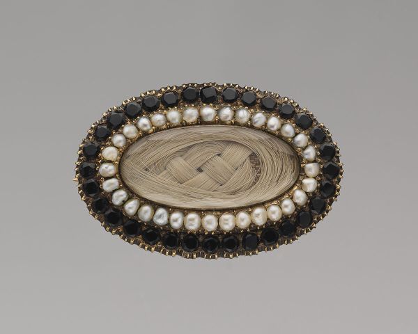 An oval brooch featuring a center of braided light brown hair, surrounded by a ring of small pearls and an outer border of dark gemstones set in gold, reflects Victorian Mourning Customs in the United States.