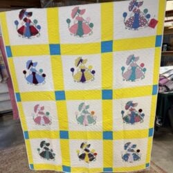 A handmade quilt featuring twelve blocks, each with an appliqué of a girl in a bonnet and dress holding an umbrella, bordered by yellow and blue sashing, displayed indoors.