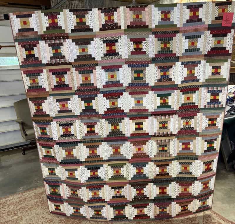 A large, colorful patchwork quilt with a geometric pattern of squares and rectangles is being held up indoors. The quilt features earthy tones, whites, and reds arranged in a repeating design.
