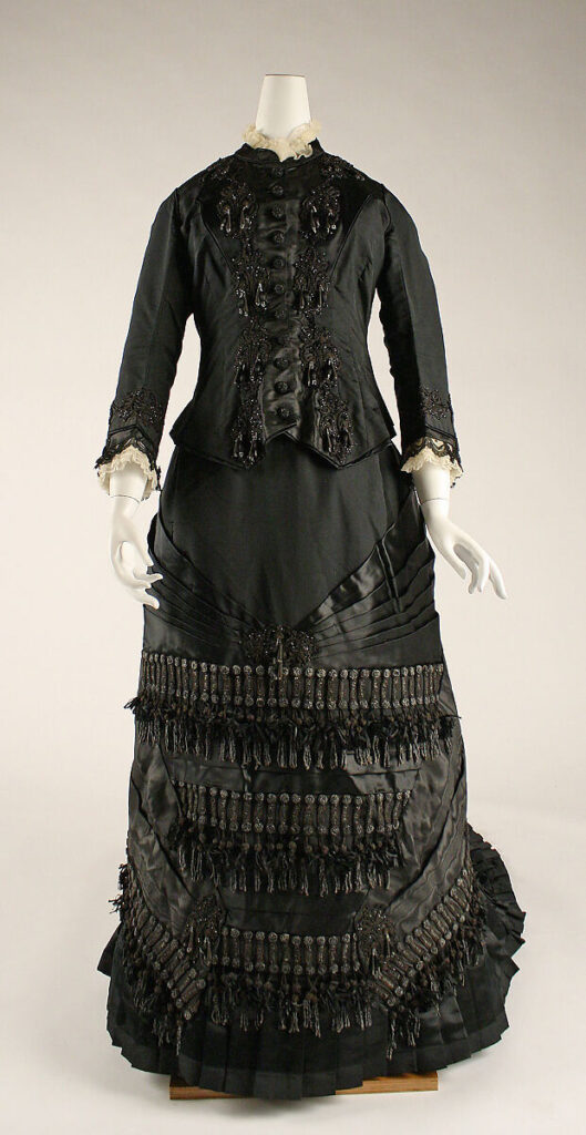 Black silk Victorian mourning dress with long sleeves, elaborate button details, and decorative tassels embodies the style of Victorian Mourning Costumes in the United States; fitted bodice features lace accents at the cuffs and collar. Displayed on a white mannequin.