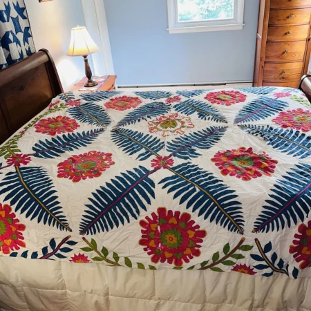 A wooden bed with a colorful quilt featuring blue fern-like leaves and pink and green floral patterns in a bright bedroom with light blue walls and wooden furniture.