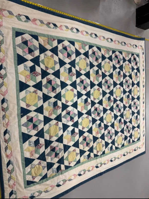 A large handmade quilt features a repeating geometric pattern with blue, yellow, and patterned fabric pieces, bordered by strips of pastel patchwork and cream fabric, displayed on a gray floor.