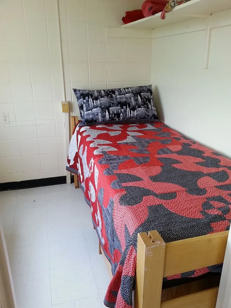 BarbM-son-college-quilt A neatly made single bed with a red, black, and gray camouflage-patterned quilt and a cityscape pillow in a small, white dorm room with a shelf above the bed.