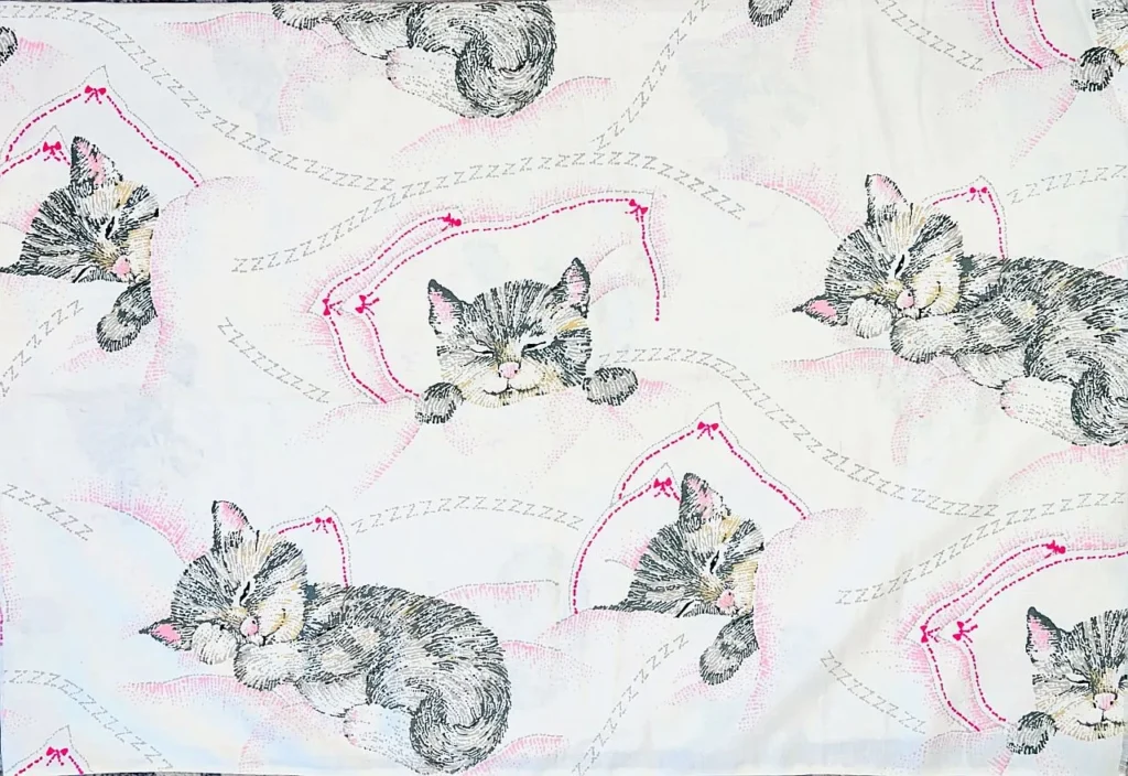 White fabric with a repeating pattern of gray tabby kittens sleeping on pink pillows, surrounded by stitched lines and “Z” shapes to indicate sleep. The design is playful and cozy, suggesting rest and comfort.
