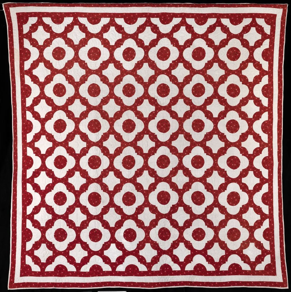 A red and white quilt featuring a repeating geometric floral pattern with red circles and petals on a white background, bordered by red and white stripes.