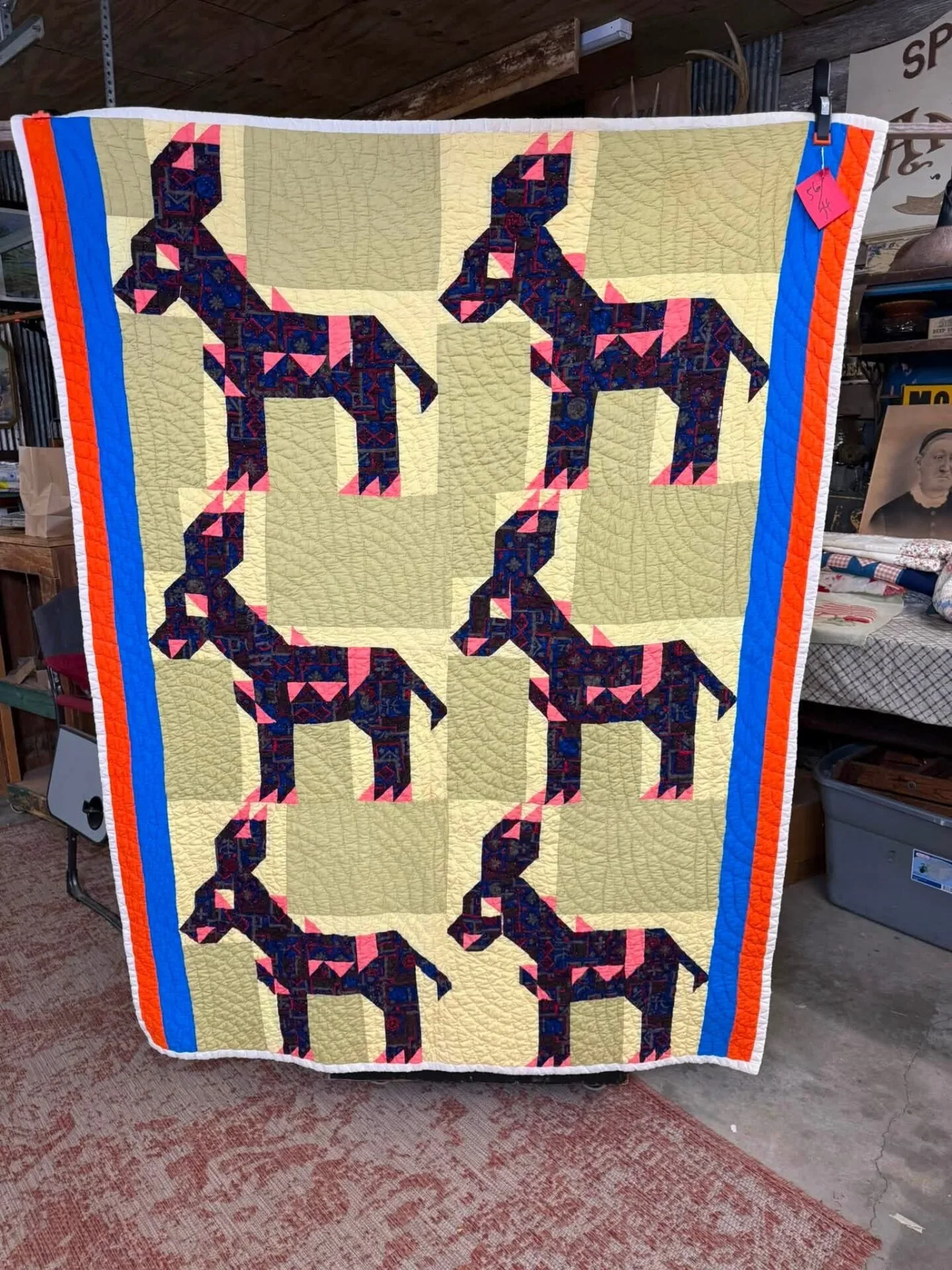 Giddyup - A handmade quilt with six patchwork donkey designs in purple and black on a yellow background, bordered by red, blue, and orange strips, hanging in a rustic indoor setting.