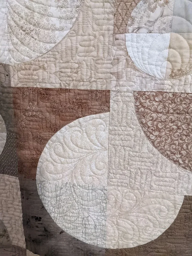 Kay-Circle-Dance A close-up of a quilt with curved and geometric patterns in beige, brown, and cream tones. The quilt features intricate stitching and various textured fabric panels.
