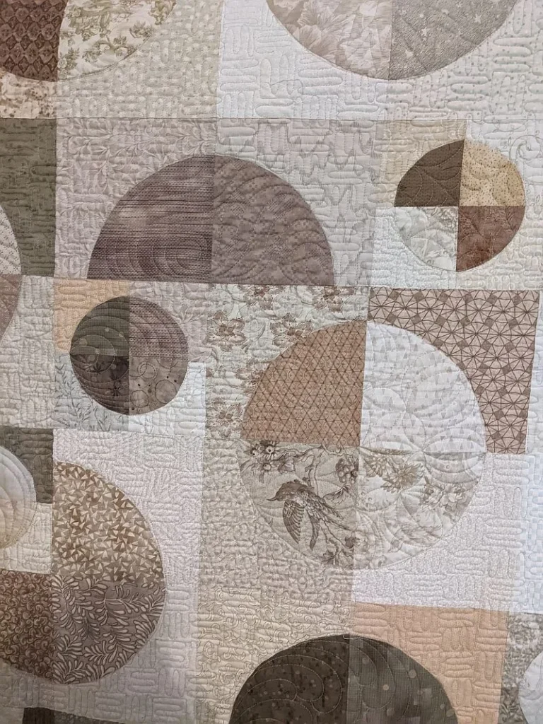 Kay-Granddaughters-wedding A quilt featuring various intersecting circles and squares in beige, cream, tan, and brown tones, with floral and geometric patterns quilted into the fabric.
