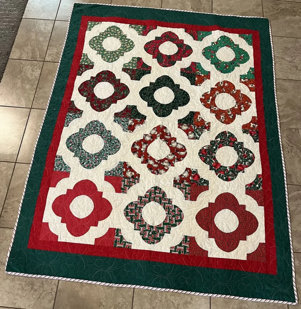 A colorful patchwork quilt with red, green, and white floral-like patterns, bordered by red and green fabric, is laid out on a tiled floor.