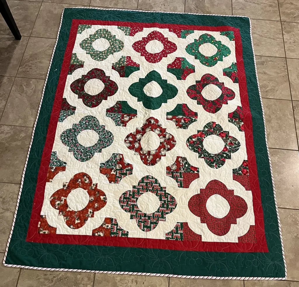 A colorful quilt with a floral pattern made of red, green, and white fabrics. The quilt has a green and red border and is placed on a tiled floor.