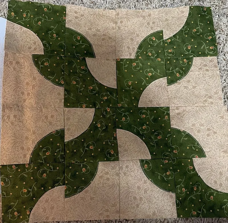 A quilt block with a curved, interlocking pattern in green and beige fabric, featuring swirling leaf-like designs on green and intricate beige prints, laid on a textured carpet background.